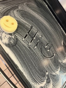 An oven door being cleaned with foam and a smiley face sponge, demonstrating a house cleaning service by Spotless by Sam LLC in Green Bay, WI.