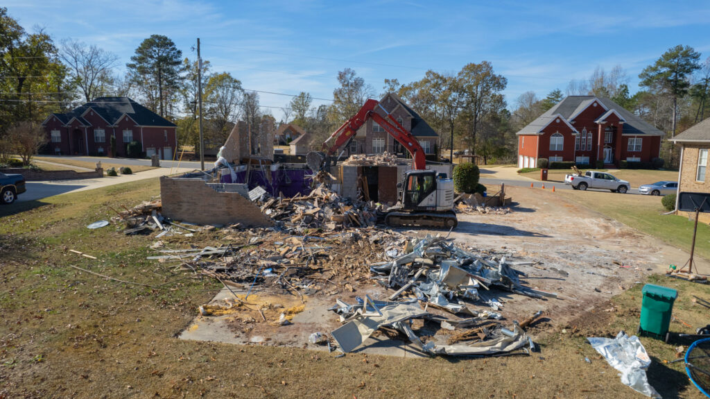 A residential house undergoing partial demolition, with an excavator visible in the background, by Nuckles Services in Birmingham, AL
