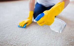 A person wearing yellow gloves cleaning a carpet with a spray bottle, demonstrating services from Home In Hand Cleaning Service in Concord, NH.