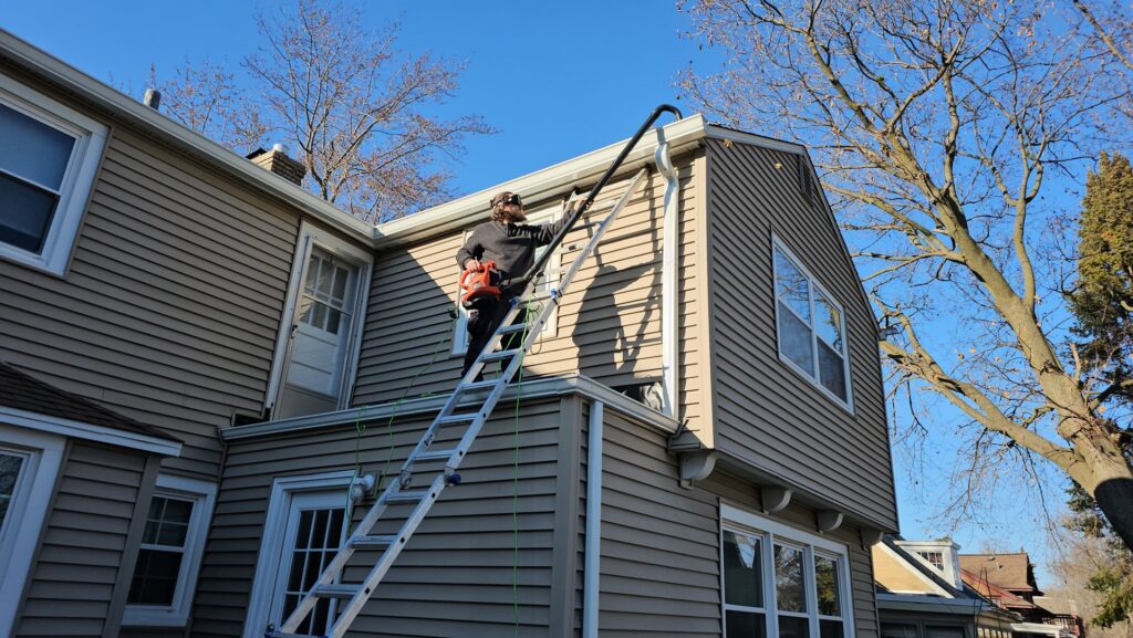 A person on a ladder using a leaf blower attachment to clean gutters on a house, a service provided by Way Too Bright Services LLC in Rockford, IL.