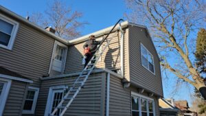 A person on a ladder using a leaf blower attachment to clean gutters on a house, a service provided by Way Too Bright Services LLC in Rockford, IL.