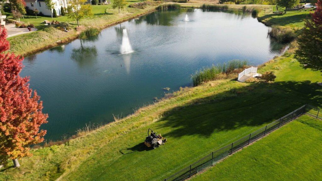 A person on a ride-on lawnmower actively cutting a large green lawn next to a pond, by Thompson Acres in Davenport, IA.