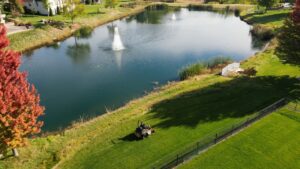 A person on a ride-on lawnmower actively cutting a large green lawn next to a pond, by Thompson Acres in Davenport, IA.