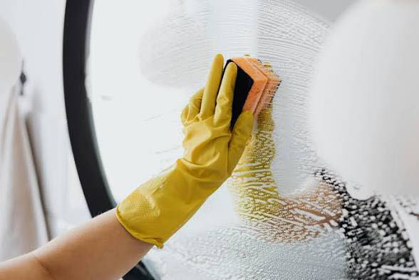 A person wearing yellow gloves scrubbing a mirror or glass surface with a sponge for Deep Cleaning Service in Frederick, MD.