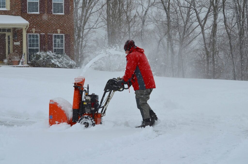 A person in a red jacket operating a snow blower to clear a snowy residential driveway for PJs Full Service Contractors in Cleveland, OH.