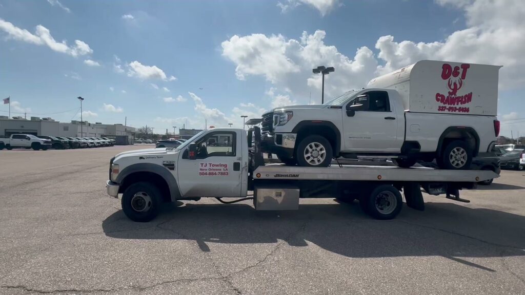 TJ Towing LLC flatbed truck towing a white pickup truck with a D&T Crawfish trailer in New Orleans, LA.