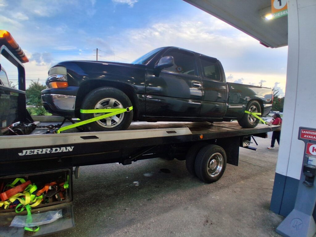 A dark pickup truck being towed on a flatbed truck by AMC Towing in Mobile, AL, at a gas station.