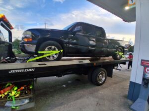 A dark pickup truck being towed on a flatbed truck by AMC Towing in Mobile, AL, at a gas station.