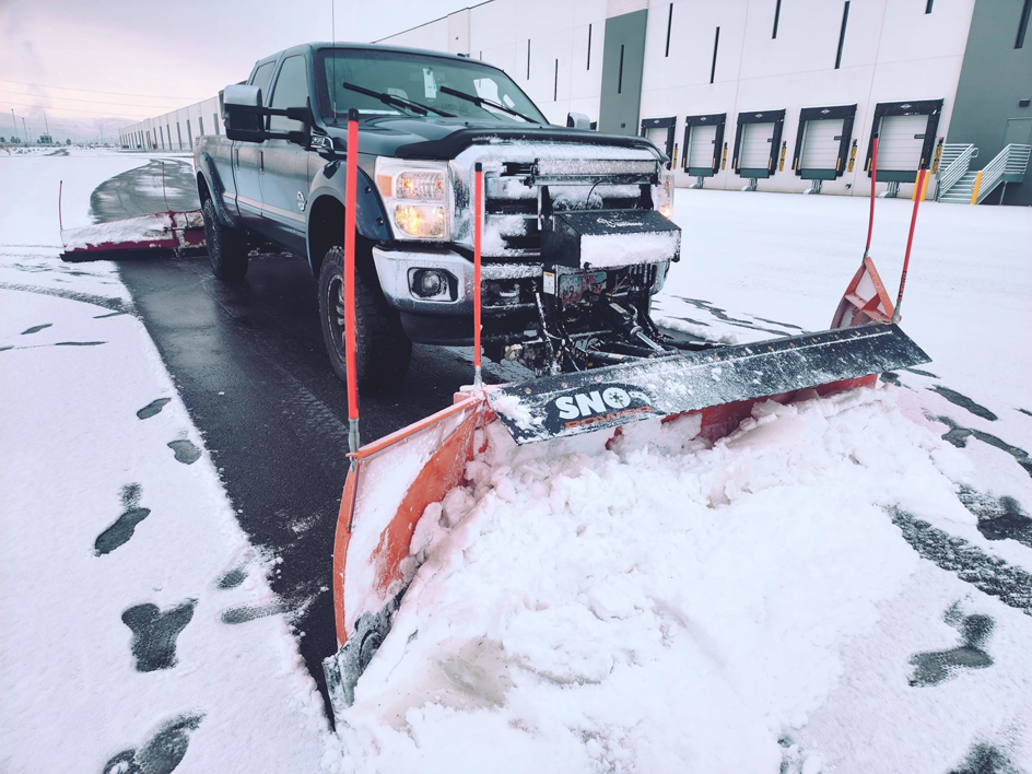 A black pickup truck with an orange snow plow actively clearing snow in a parking lot for Mr Plow in South Salt Lake, UT.