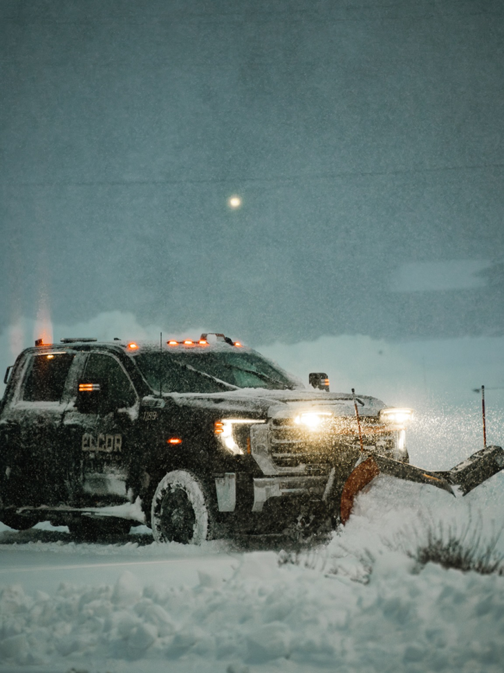 A black pickup truck with a snowplow attachment clearing snow on a road for Elcor Construction in Rochester, MN.
