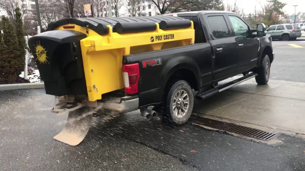 A pickup truck with a Poly Caster salt spreader actively spreading salt on a paved surface for JC Snow Management in Mount Vernon, NY
