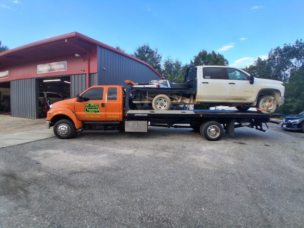 An AMC Towing flatbed truck in Mobile, AL, transporting a white pickup truck to a repair shop.