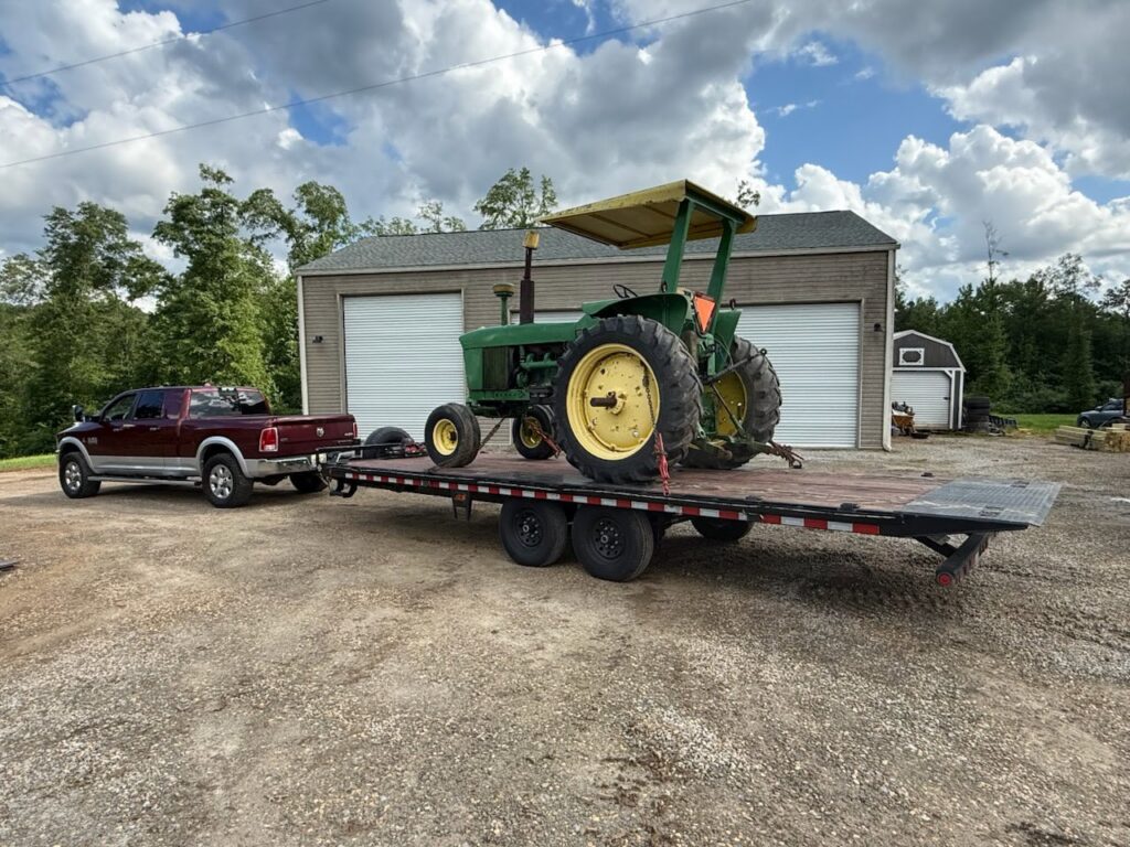 A pickup truck towing a large green tractor on a flatbed trailer from B'ham Trailer Rentals in Alabaster, AL.