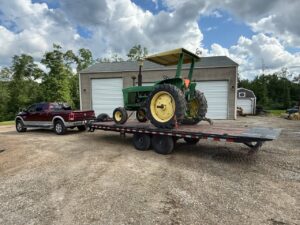 A pickup truck towing a large green tractor on a flatbed trailer from B'ham Trailer Rentals in Alabaster, AL.