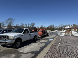 A white pickup truck towing a Kubota skid steer and other equipment in a snowy parking lot for Hays Landscape Solutions LLC in Jackson, TN.