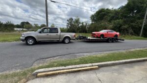 A pickup truck towing an orange sports car on a flatbed trailer from B'ham Trailer Rentals in Alabaster, AL.