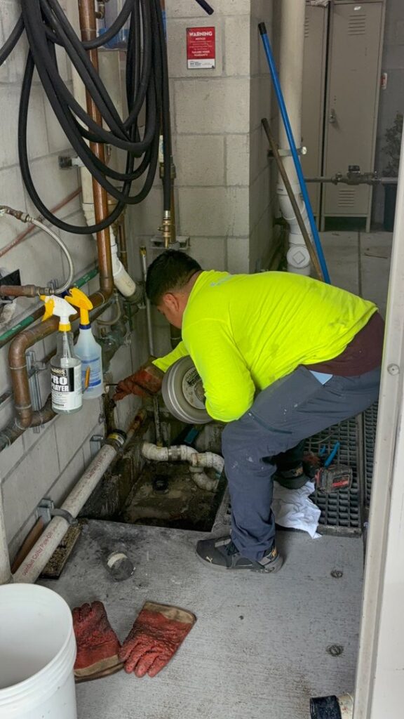 A Z and Z Plumbing specialist working on drain pipes near a floor drain in a utility room in Oakland, CA.