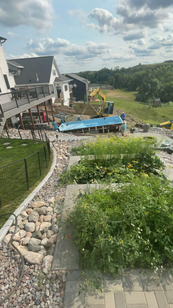 A swimming pool shell being carefully lowered into an excavated hole during installation by Backyard Escapes by Kerner Pools & Spas in Bismarck, ND.