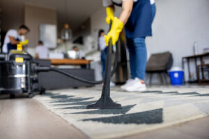 A professional cleaner in yellow gloves vacuuming a carpet, demonstrating a house cleaning service by Peoria Cleaning Pros, LLC in East Peoria, IL.