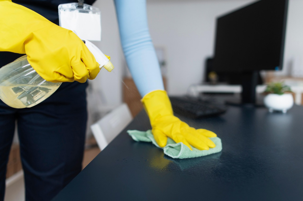 A professional cleaner wearing yellow gloves wiping a desk surface for Ready.Set.Clean LLC in Morley, MI.