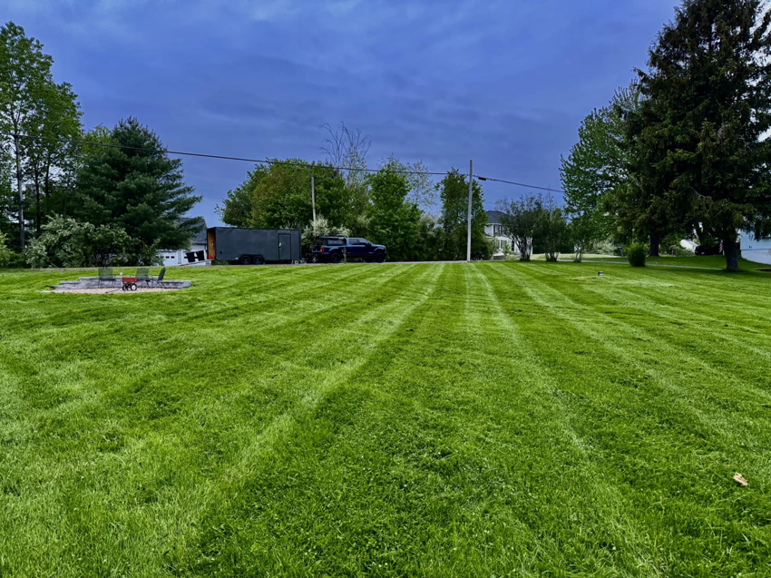 A large, professionally mowed lawn with distinct stripes and landscaping equipment in the background by Merrily Landscaping LLC in Derry, NH.