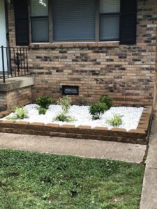 A beautifully designed raised garden bed with white gravel and small plants in front of a brick house by The Otherside Lawn Solutions in Ankeny, IA.