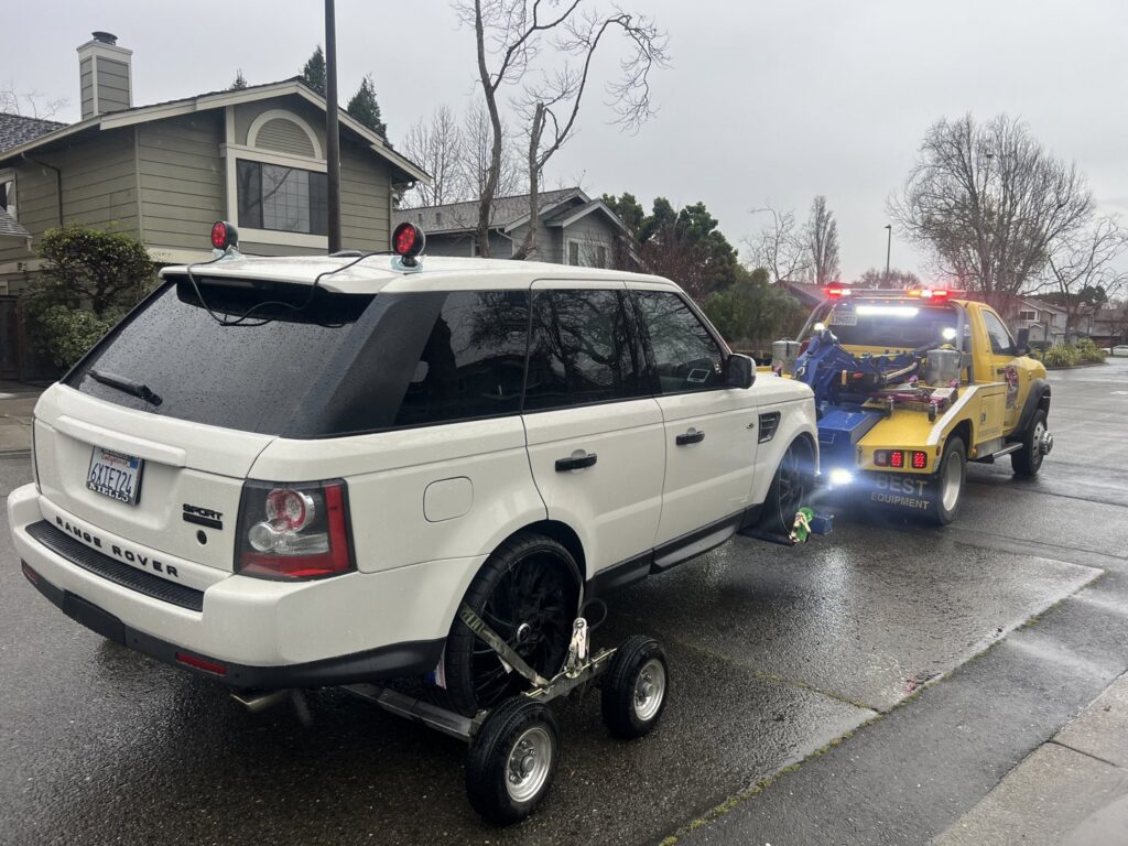 A white Range Rover SUV is being towed by a wheel-lift tow truck from S&F Towing in Oakland, CA.