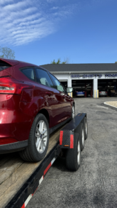 A red car being towed on a flatbed trailer in front of an auto shop by 82Hauling & More LLC in St. Louis, MO