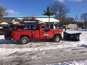 A red pickup truck equipped with a snow plow parked in a snowy commercial lot by Haynes Snow Removal & Lawn Maintenance in Madison, WI.