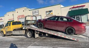 A red sedan being loaded onto a flatbed tow truck by Geely Towing outside an auto repair shop in Fargo, ND.