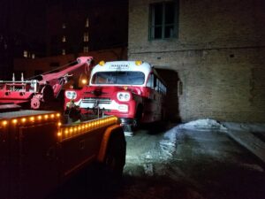 A red tow truck from Chicago Towing towing a vintage red and white bus in the dark in Chicago, IL.