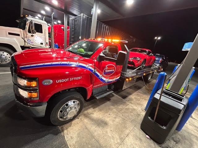 A red flatbed tow truck from Advance Towing & Recovery transporting a red sports car at night in Columbus, GA.