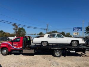A red flatbed tow truck from Advance Towing & Recovery transporting a white classic sedan in Columbus, GA.