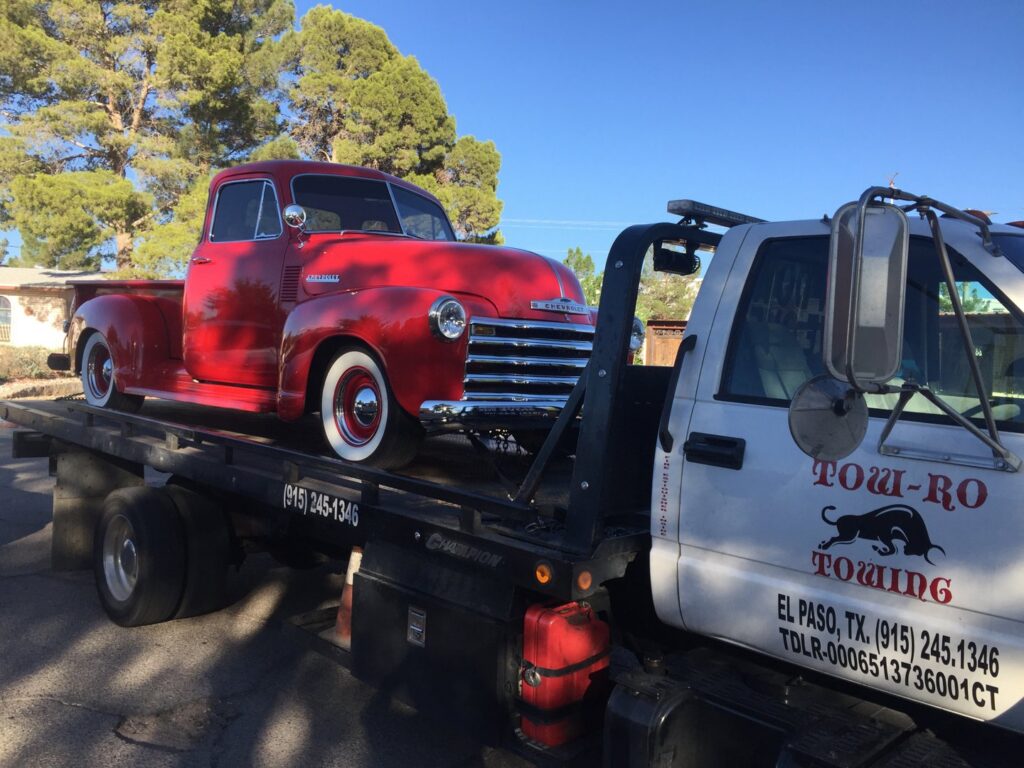 A red vintage pickup truck being transported on a flatbed tow truck by Tow-Ro Towing in El Paso, TX.