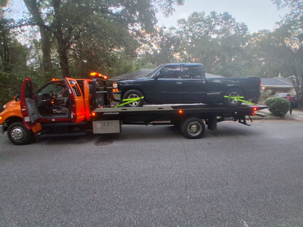 A dark pickup truck being towed on a flatbed truck by AMC Towing in Mobile, AL, on a residential street.