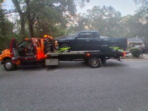 A dark pickup truck being towed on a flatbed truck by AMC Towing in Mobile, AL, on a residential street.