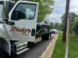 Ricky's Towing & Recovery LLC flatbed tow truck transporting a black car, with a police vehicle in the background, performing a service in Derwood, MD.
