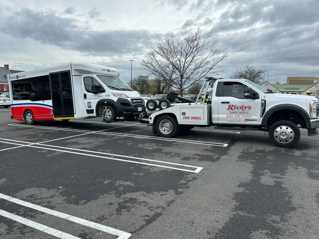 A Ricky's Towing & Recovery LLC tow truck using a wheel lift to tow a Metro Access bus in a parking lot, providing service in Derwood, MD.