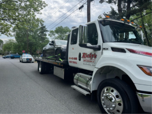 A Ricky's Towing & Recovery LLC flatbed truck actively transporting a black sedan, with a police car in the distance, providing service in Derwood, MD.