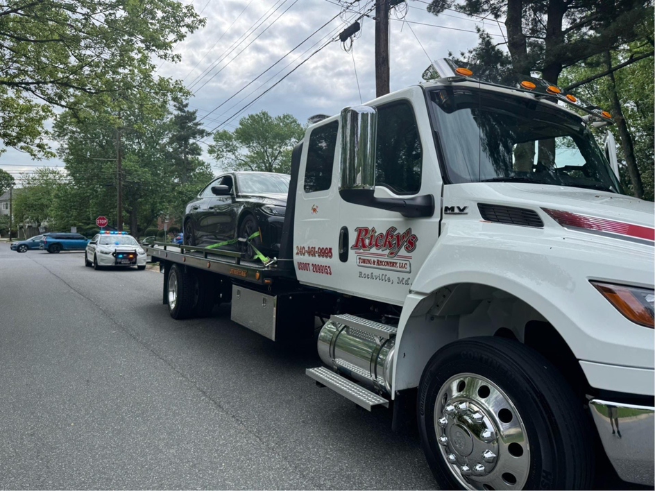 A Ricky's Towing & Recovery LLC flatbed truck actively transporting a black sedan, with a police car in the distance, providing service in Derwood, MD.