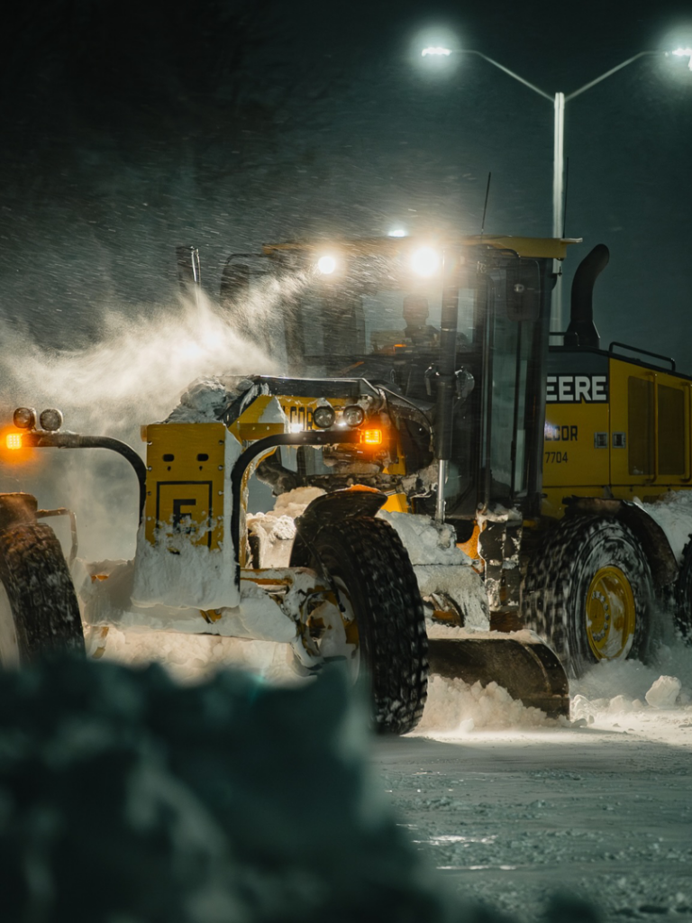 A yellow John Deere road grader actively plowing snow at night for Elcor Construction in Rochester, MN.