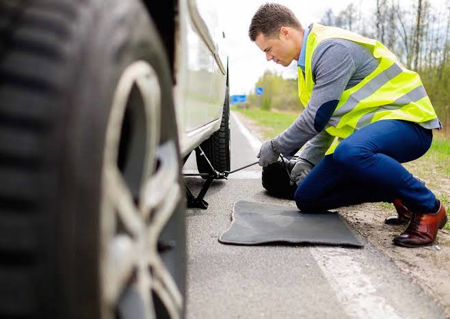 A service technician in a high-visibility vest changing a flat tire on the side of the road, a service provided by Emergency Towing Sacramento, CA.