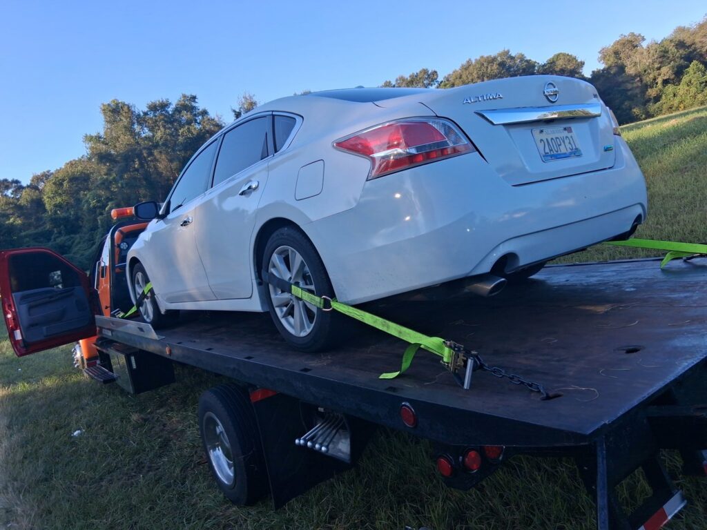 A white Nissan Altima being towed on a flatbed truck by AMC Towing in Mobile, AL, from the side of a road.