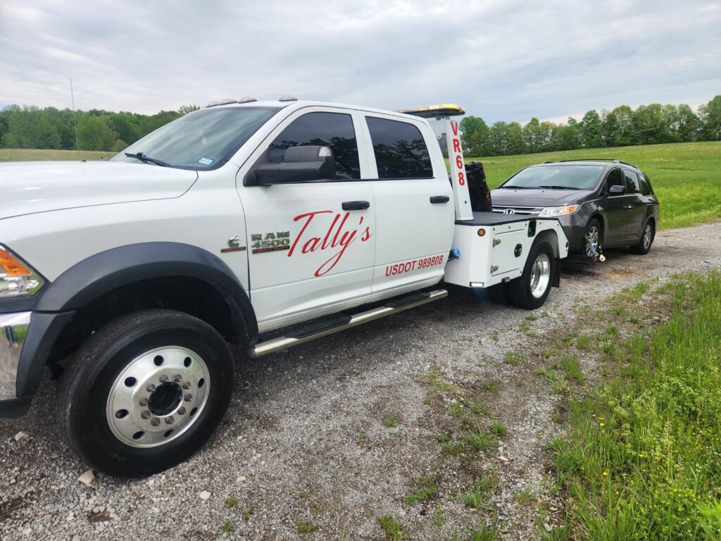 A white rollback tow truck from Tally's Towing and Recovery towing a dark SUV on a dirt road in Owensboro, KY.