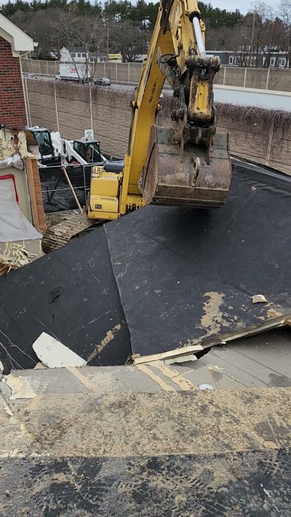 An excavator performing roof demolition on a building, with another machine in the background, by Allied Wrecking Boston in East Boston, MA.