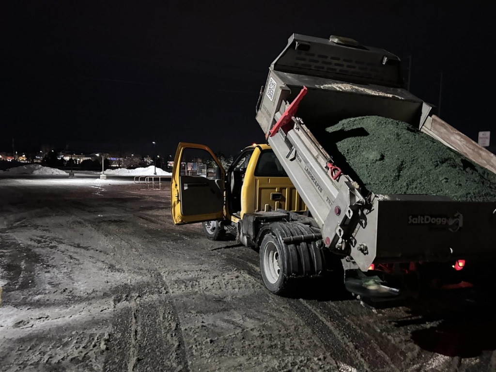 A dump truck with a SaltDogg spreader applying de-icing salt to a snowy parking lot at night by Cities Lawn and Snow LLC in Woodbury, MN.