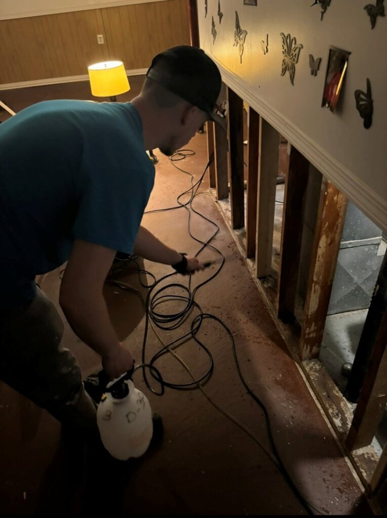 A water damage specialist spraying a sanitizing solution on a water-damaged floor with exposed studs during remediation by H2O Pros Water Restoration Services in Westland, MI.