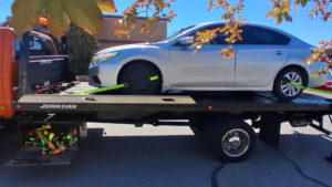 A silver sedan being towed on a flatbed truck by AMC Towing in Mobile, AL, with tree branches in the foreground.