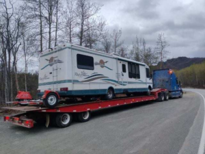 A semi-truck from Help Line Towing transporting a large RV on a flatbed trailer on a road in Wasilla, AK.