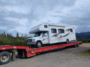 A semi-truck from Help Line Towing transporting an RV on a flatbed trailer under a cloudy sky in Wasilla, AK.